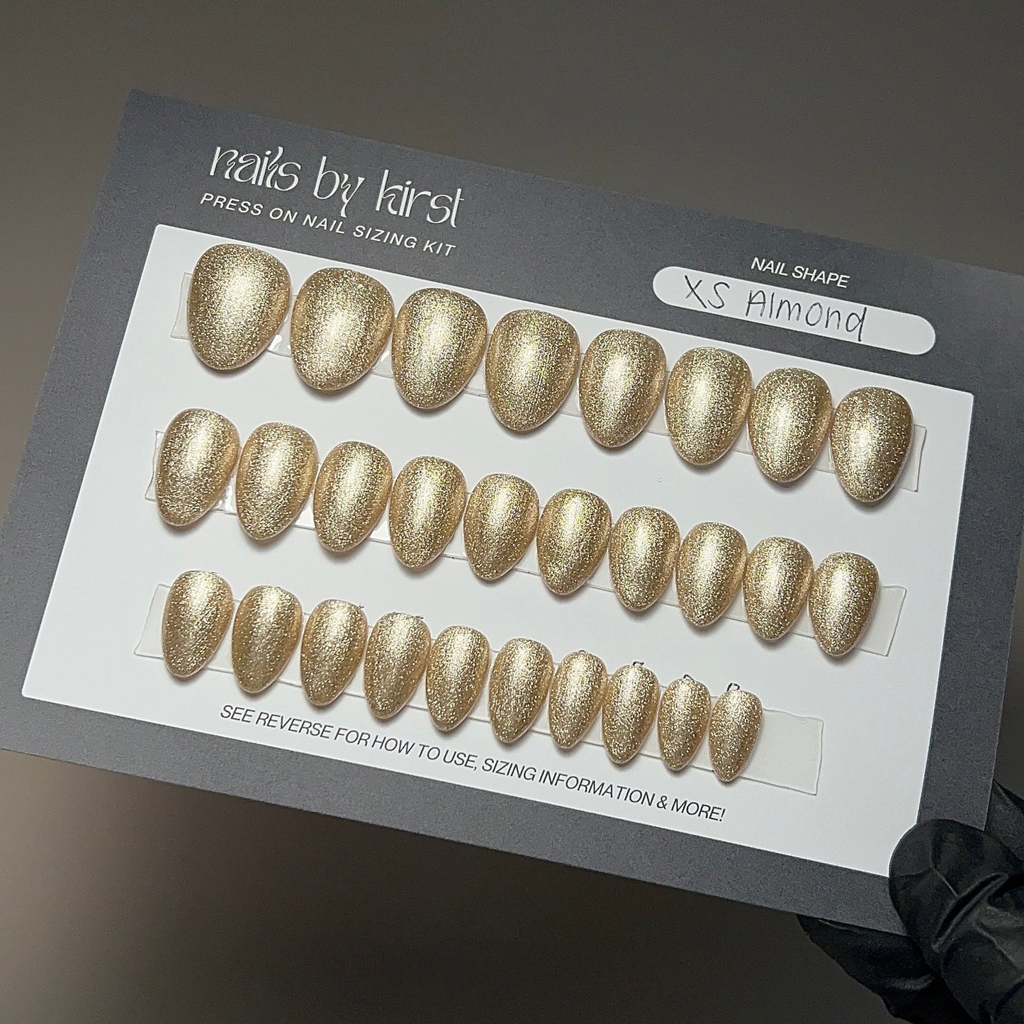 Gold press-on nail set on a gray card with 'Nails by Lirst' branding.