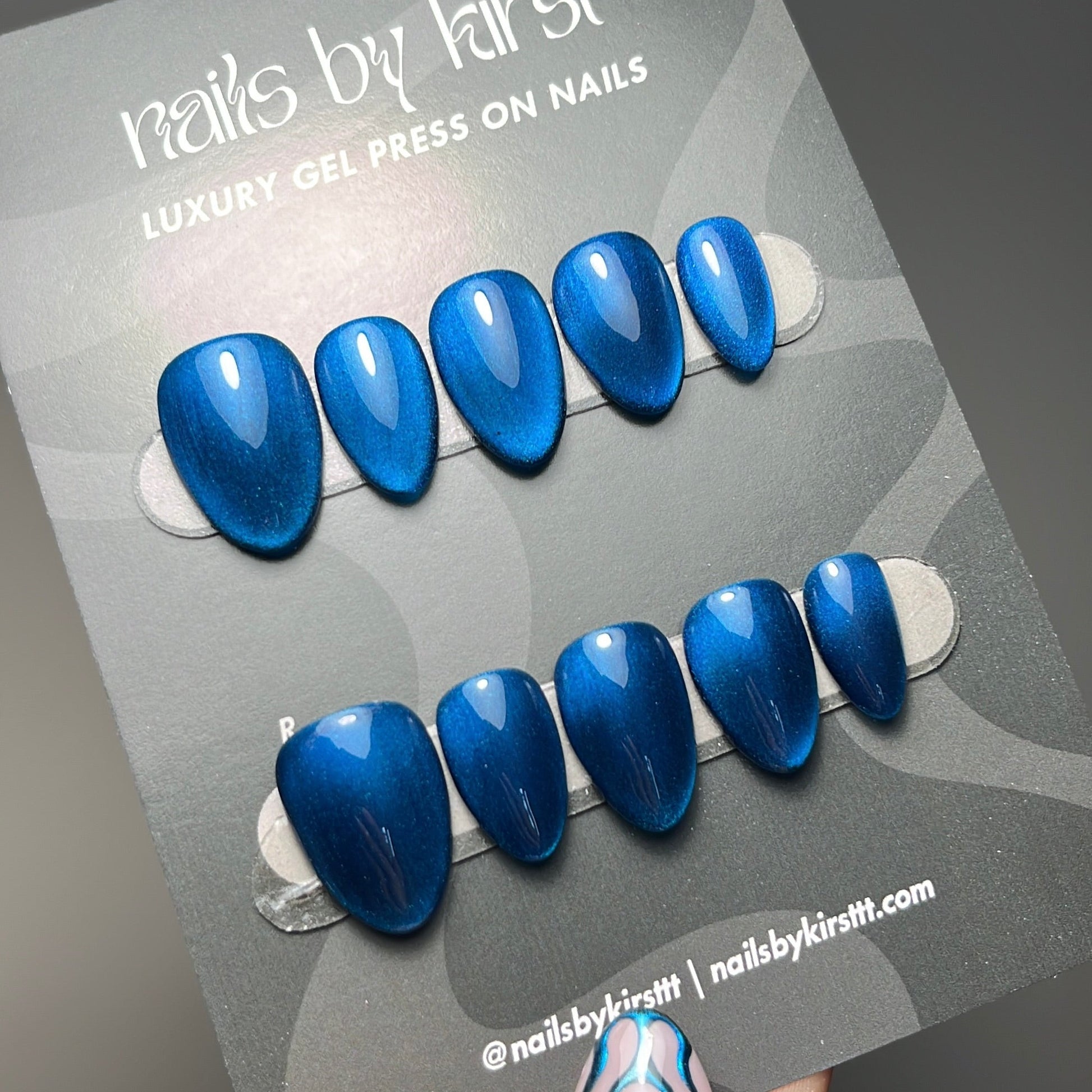 A hand holding a card of blue magnetic press-on nails. The nails have a glossy metallic blue finish with a subtle cat-eye effect, and the card is branded 'nails by kirst'.
