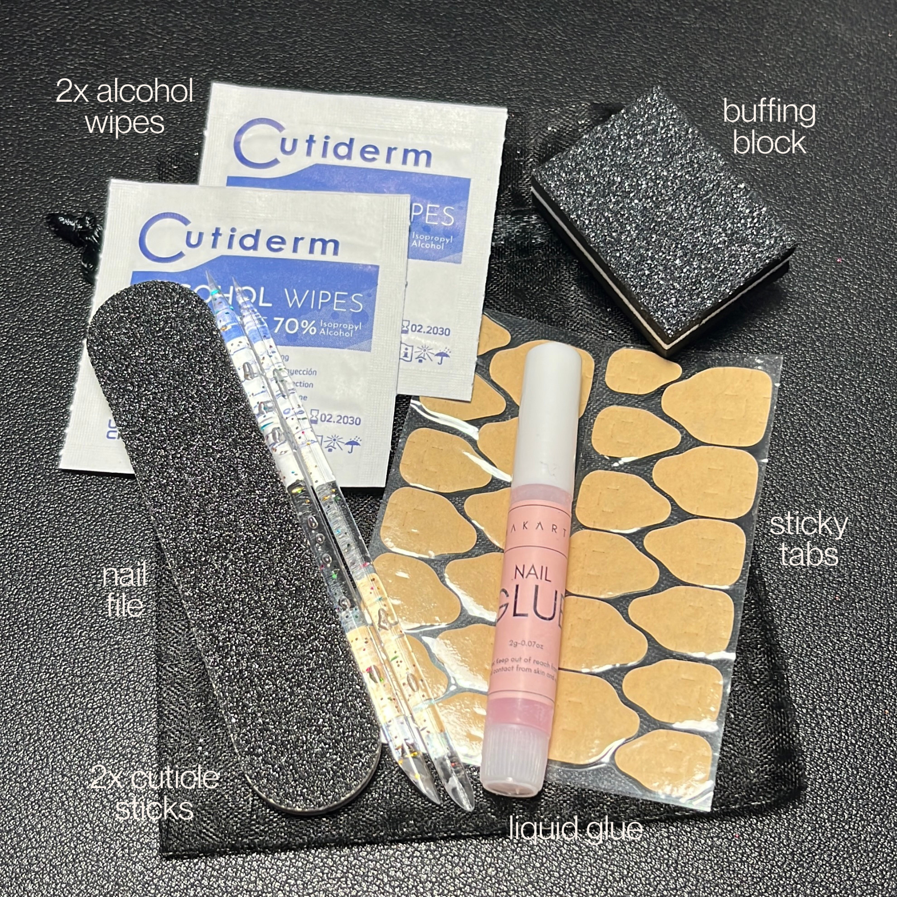 Collection of nail care items including alcohol wipes, cuticle sticks, a buffer block, nail glue, and sticky tabs on a black surface.