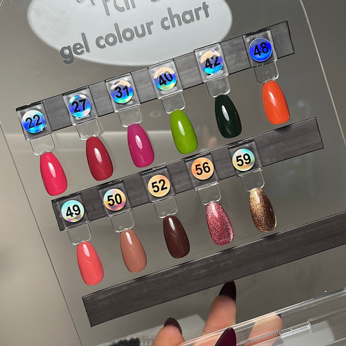 A hand holds a clear acrylic nail color chart display labeled 'nails by kirst gel colour chart' with multiple gel nail swatches in different colors 