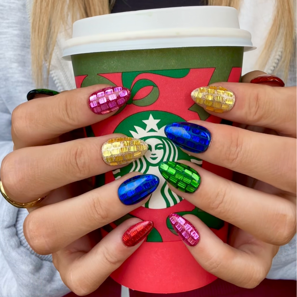 A close-up of a person holding a Starbucks coffee cup, showcasing vibrant, multicolored nail art with gold glitter, blue, green, pink, and red designs.