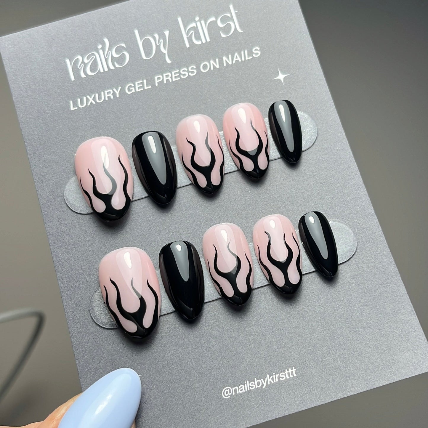 Set of press-on nails with black flame design on a gray card, held by a hand with light blue nail polish.