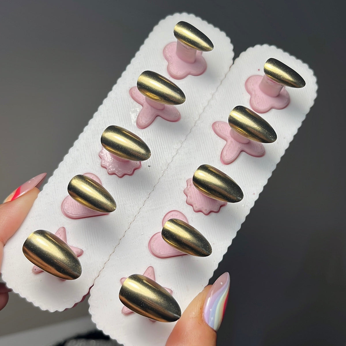 A hand holding two white nail display cards with metallic gold almond-shaped press-on nails, set against a dark gray background.