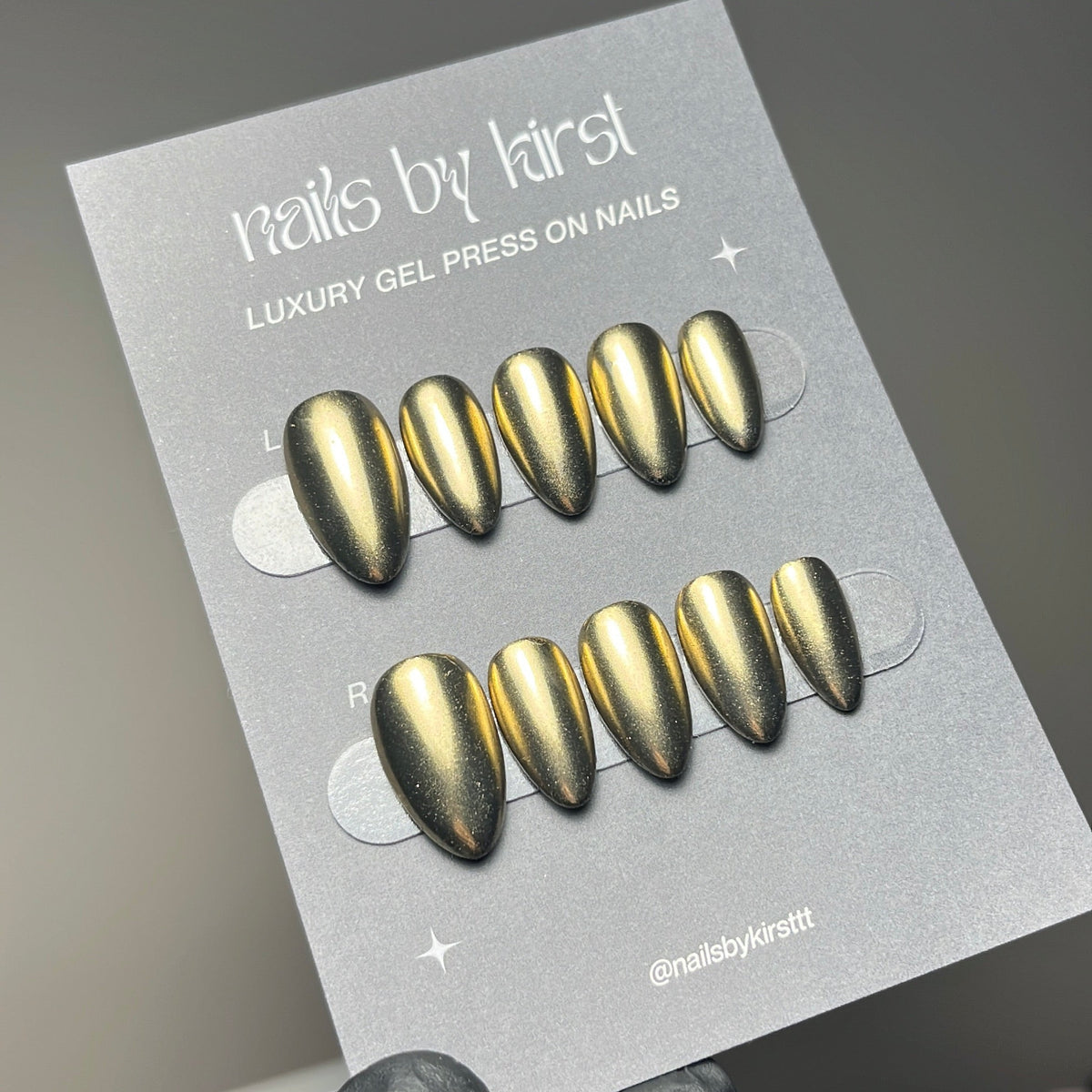 A hand wearing a black glove holds a card displaying metallic gold press-on nails. The card reads 'nails by kirst' and 'LUXURY GEL PRESS ON NAILS'.