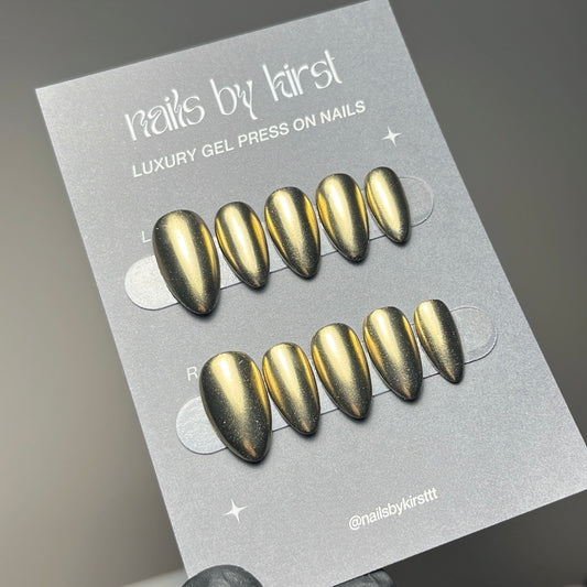A hand wearing a black glove holds a card displaying metallic gold press-on nails. The card reads 'nails by kirst' and 'LUXURY GEL PRESS ON NAILS'.