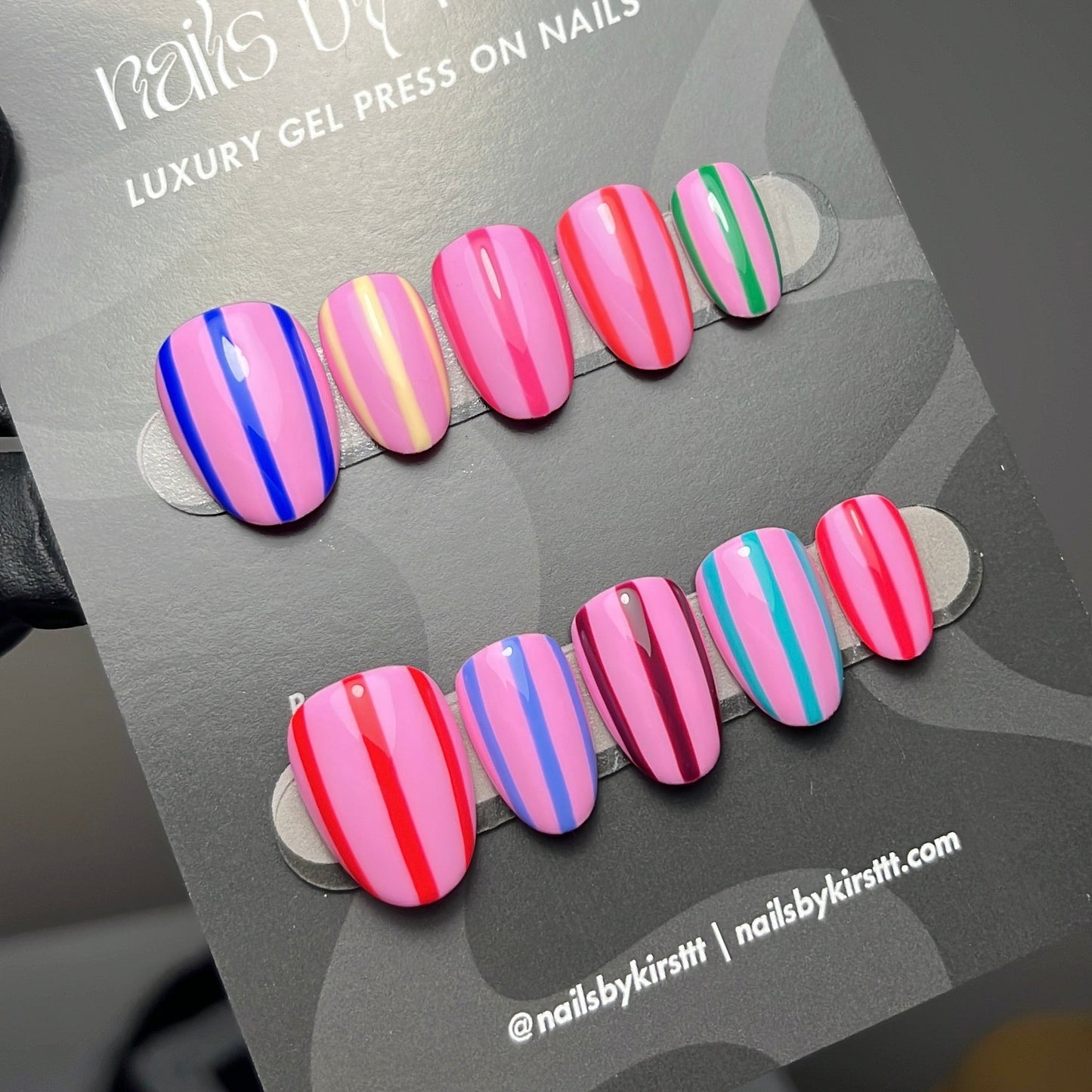 A set of artificial press-on nails displayed on a gray card labeled 'nails by kirst'. The nails feature stripe patterns.