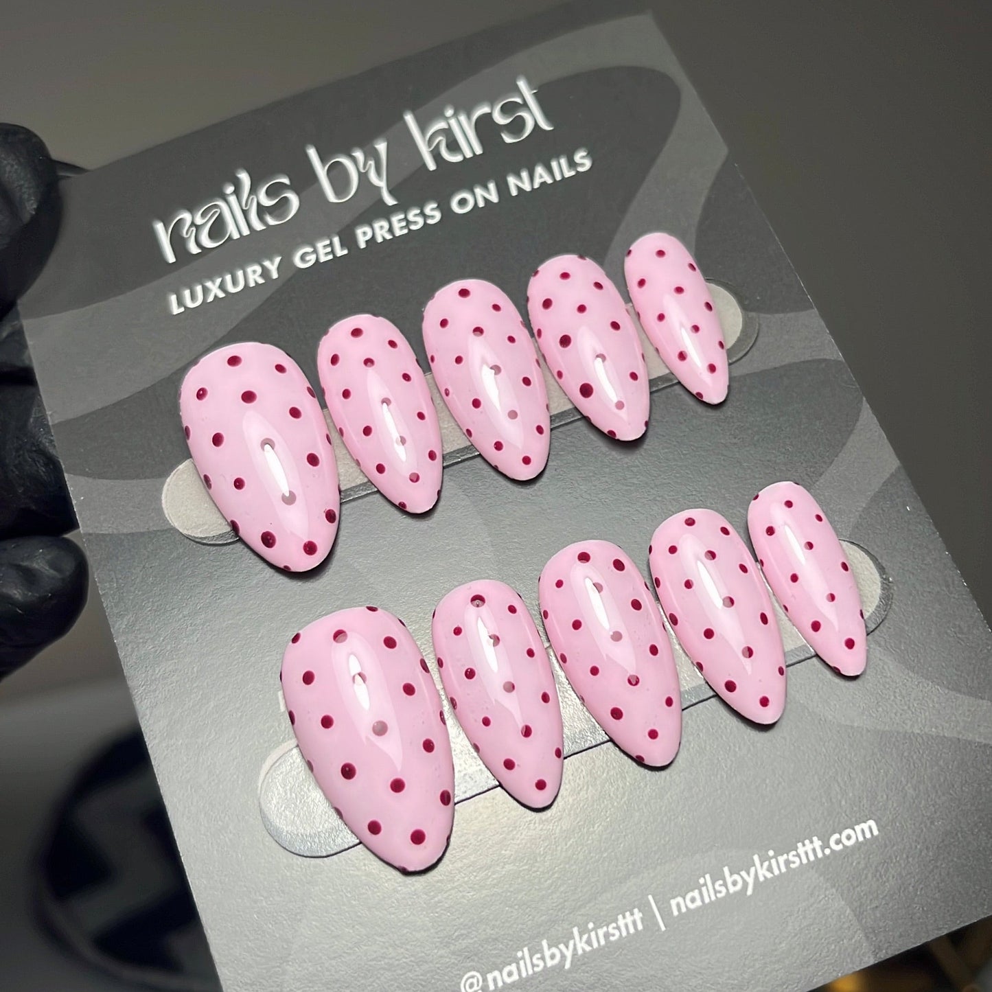 Pink press-on nails with dark pink polka dots on a gray display card, held by a hand wearing a black glove. The card reads 'nails by kirsttt' and 'LUXURY GEL PRESS ON NAILS'.