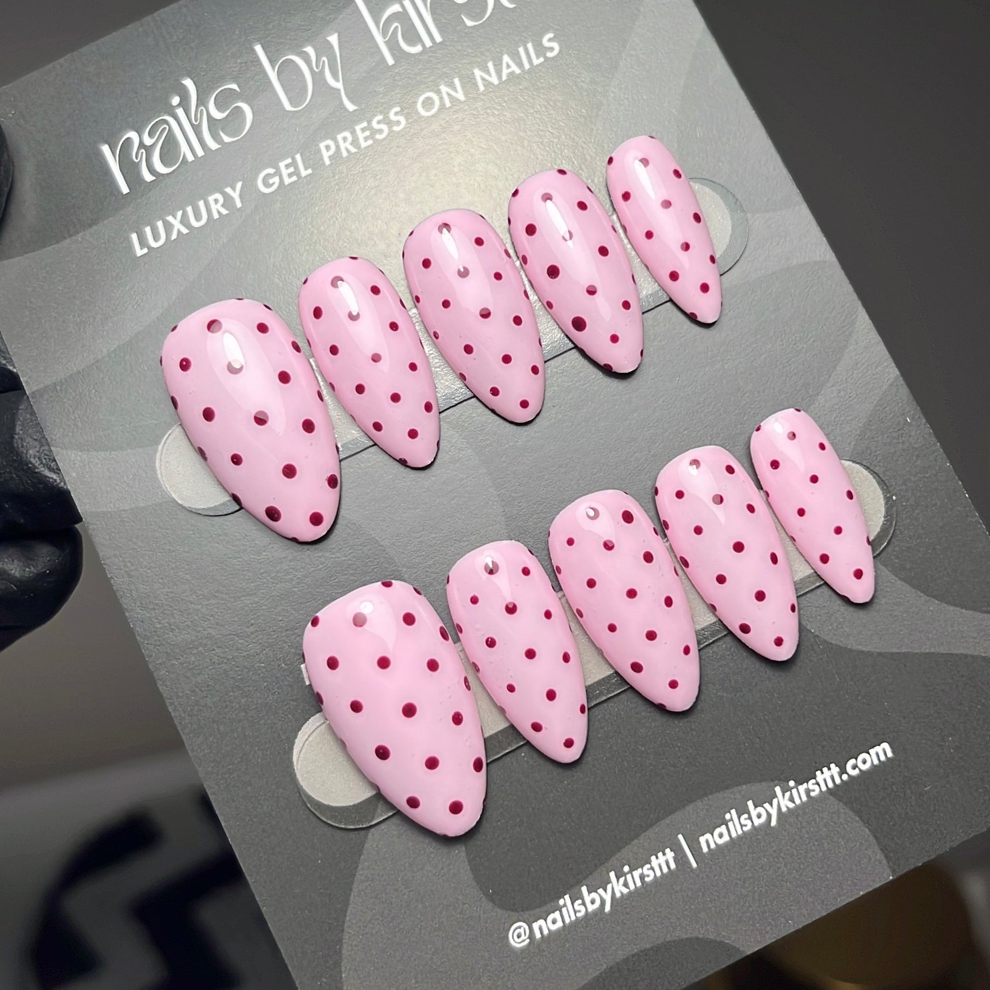 A set of pink press-on nails with small red polka dots, displayed on a gray card labeled 'nails by kirsttt'.