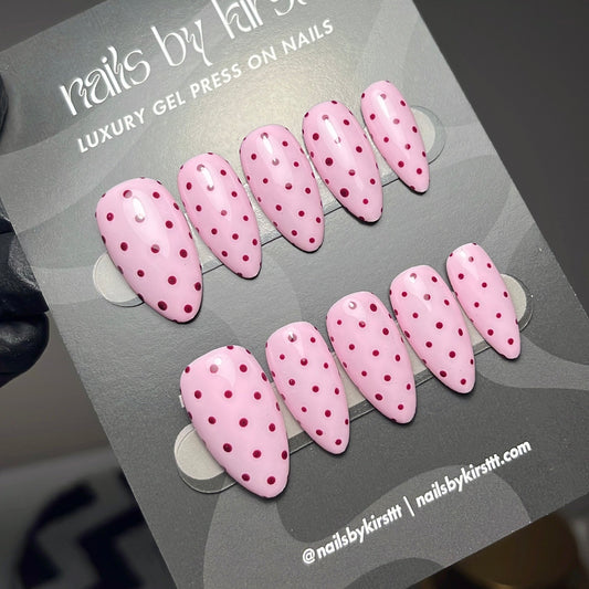 A set of pink press-on nails with small red polka dots, displayed on a gray card labeled 'nails by kirsttt'.