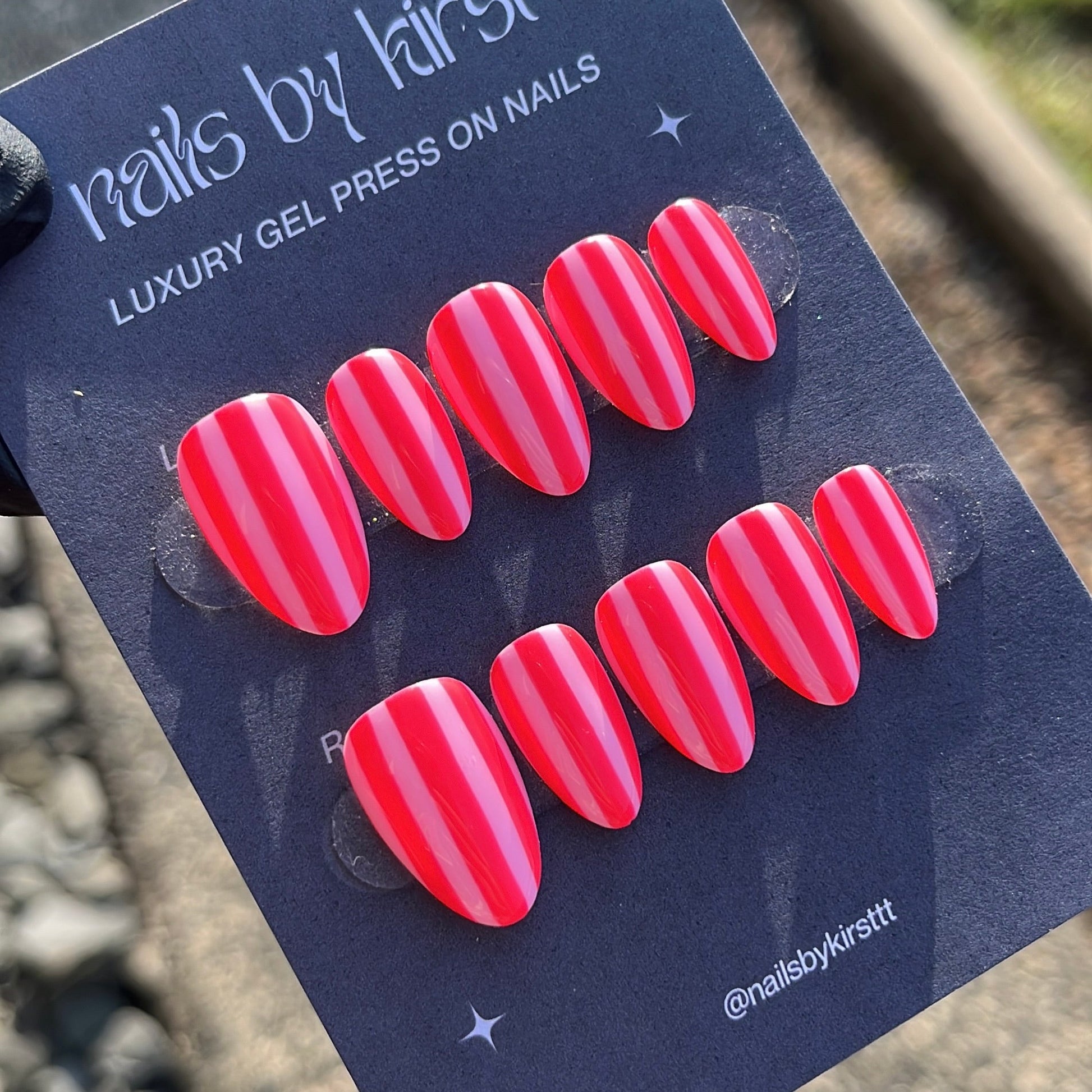 Red gel press-on nails on a 'nails by kirst' branded card against a blurred outdoor background.