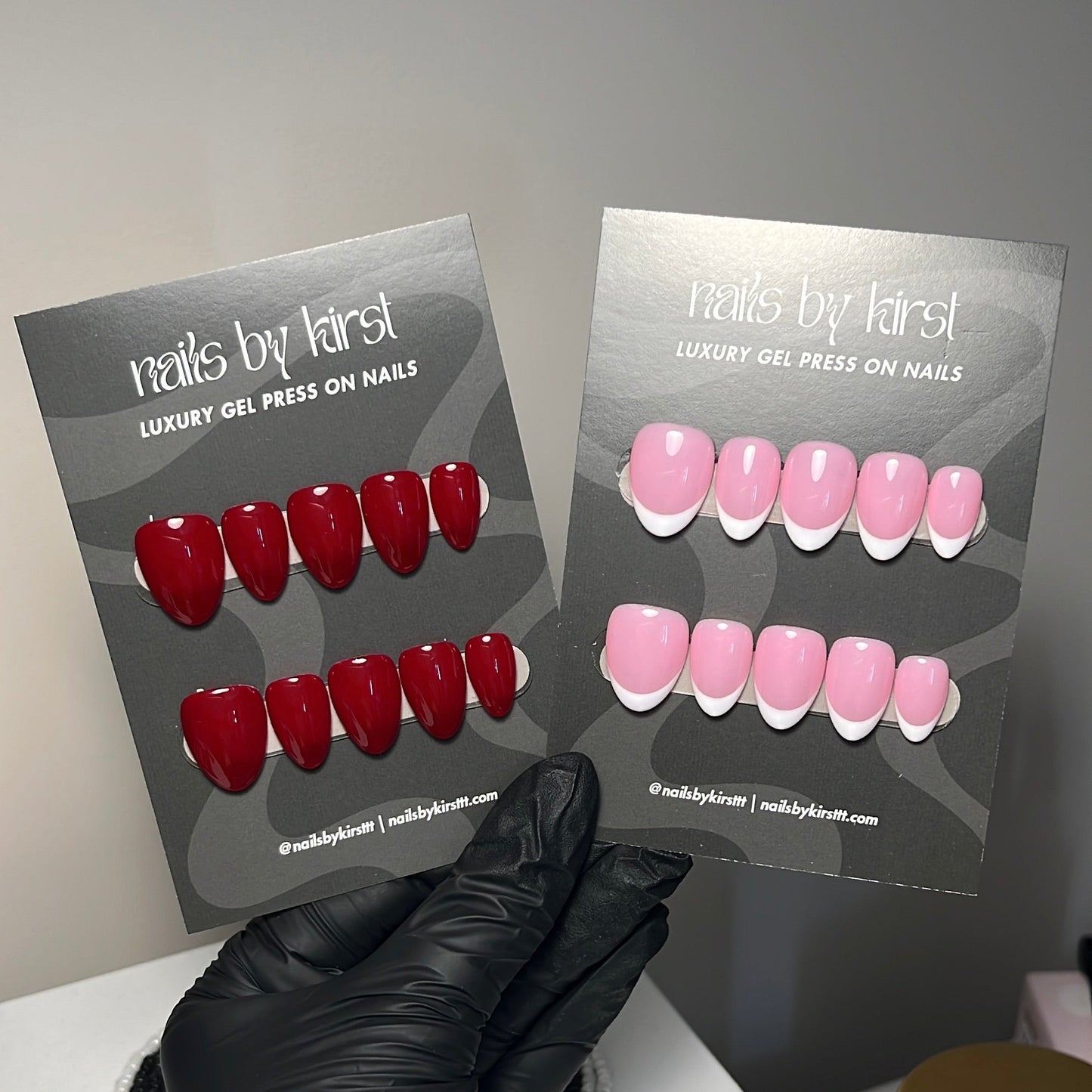 A hand wearing a black nitrile glove holds two display cards showing press-on nails. The left card displays glossy red press-on nails, and the right card displays light pink press-on nails with white french tips.