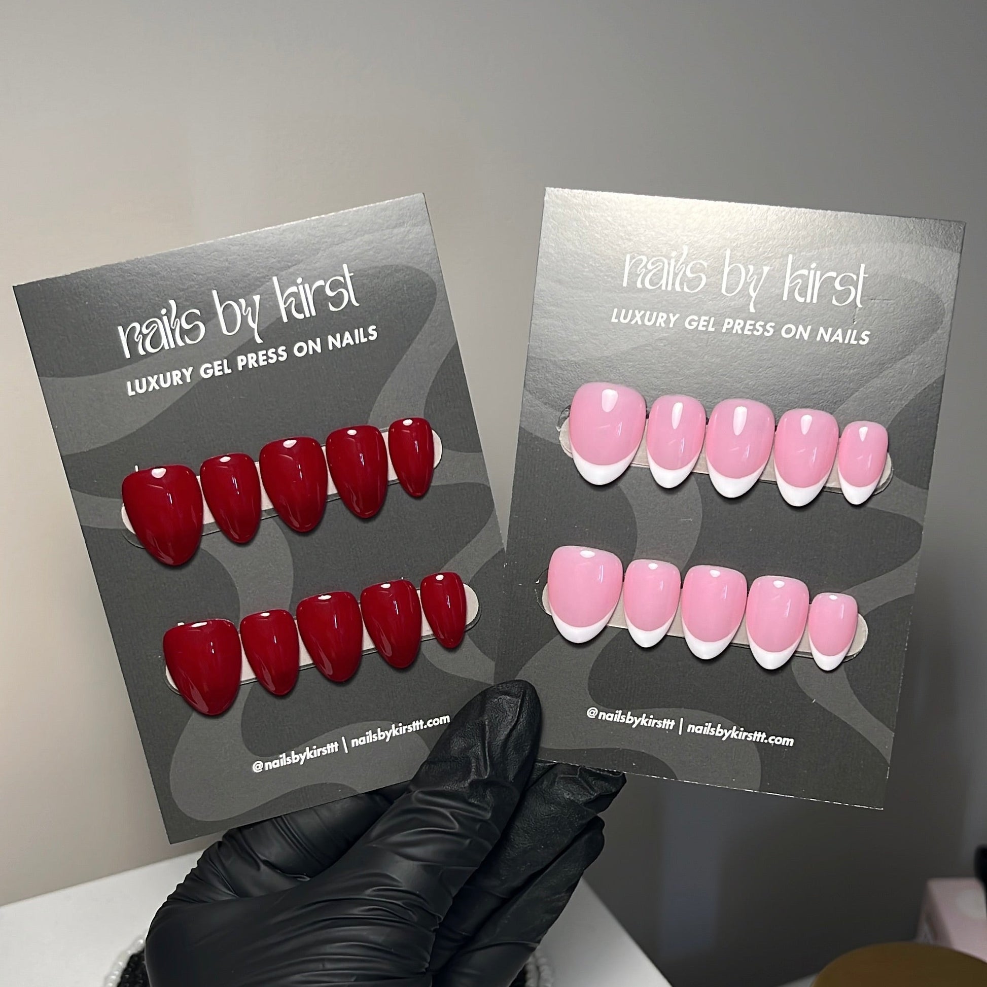 A hand wearing a black nitrile glove holds two display cards showing press-on nails. The left card displays glossy red press-on nails, and the right card displays light pink press-on nails with white french tips.