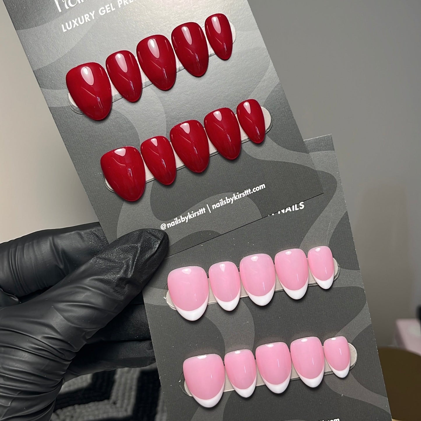 A hand wearing a black glove holds a display card showing two sets of press-on nails: glossy red nails on the top and glossy pink nails with white french tips on the bottom. The card reads 'nails by kirst' and includes social media text.